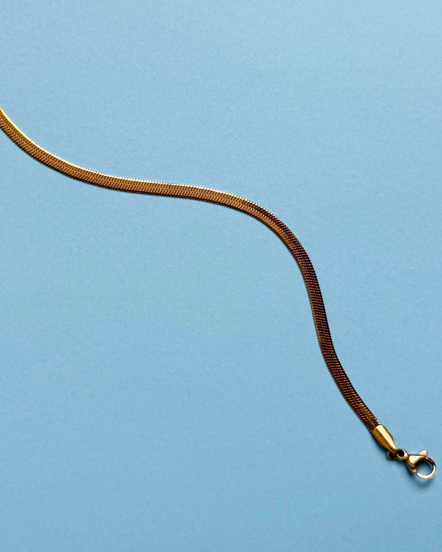 A single long, thin gold herringbone bracelet, photographed on a smooth light blue background.